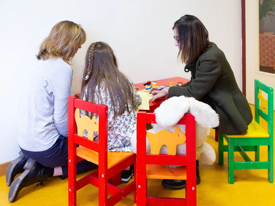Une fille et deux femmes adultes sont assises à une petite table, vues de dos.