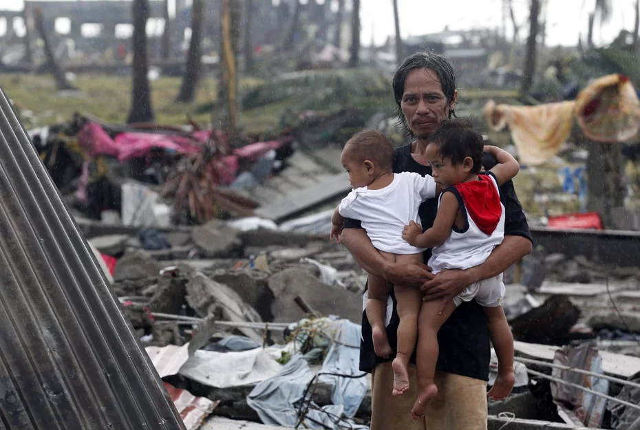 Un homme sourit fermement en tenant deux enfants au milieu des destructions causées par le typhon Haiyan en 2013, faisant preuve de résilience et d'espoir.