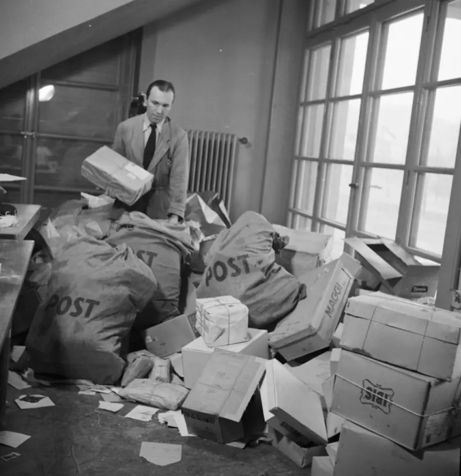 Un homme trie de grands sacs de lettres et de paquets, symbolisant les premiers efforts internationaux de la Chaîne du Bonheur après la guerre.